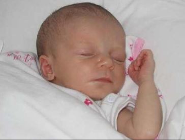 Tessa is een pasgeboren baby, ze ligt te slapen in een bedje met witte lakentjes, een handje naast haar hoofd. Ze heeft dunne babyhaartjes. 