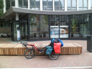 Ligfiets, zwaar bepakt met grote fietstassen voor een gebouw. Door het raam heen is een hele snelle ligfiets te zien met een aerodynamische overkapping.  