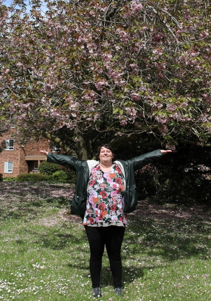 Rosie in front of a tree full of pink blossom. She stands with her arms wide and wears a matching top with all flowers in red, pink, white and black, a dark green coat and black pants. 