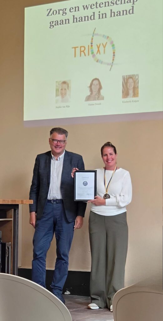 Sophie kreeg de Betto Deelman prijs. Op de foto met de oorkonde. Naast haar dr Alex Knipping. Op de achtergrond een dia met de tekst 'Zorg en wetenschap gaan hand in hand'